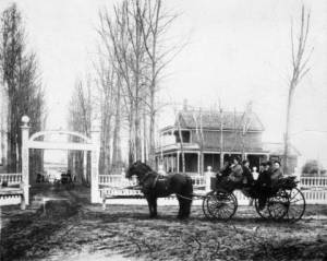 Home of Mr. Addison Nathan Baker on Grand Avenue (West Colfax Avenue) in Denver, Colorado. The farm house is a two story brick structure surrounded by a fence with a gate. The Baker family, men, women, and children, sit in a horse drawn carriage.