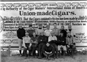 Members of the Denver Bears baseball team pose outdoors at a baseball field in Denver, Colorado. Some of the men wear uniforms. A cigar union sign is used as the backdrop. Courtesy History Colorado Collection CHS-B1369