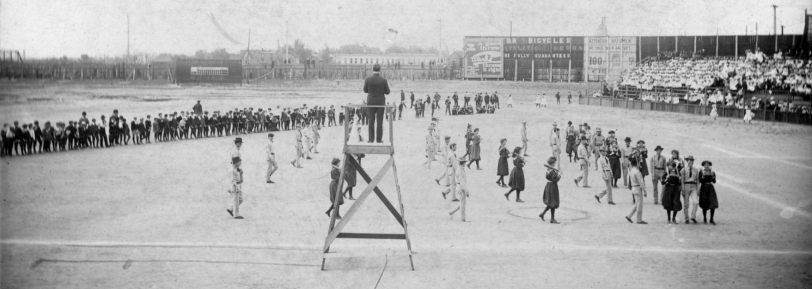 Broadway Park II. Men and women perform an exercise demonstration at the athletic field in Broadway Park, West 6th (Sixth) Avenue and Broadway in Denver, Colorado. A man stands on a wooden elevated platform and oversees the athletes. The women, in bloomers, hold Indian clubs, and march with men in suits, ties and cowboy hats who carry exercise sticks. Men and women stand near the field or sit on a fence or on bleachers. Painted signs read: "Dr. Bicycles Athletic Goods, fully guaranteed" "Earle, a good throw throws it right" and "Attention batsmen, hit this sign and get 100 US 5 [cents] Ci[gars]." [between 1900 and 1920?] Courtesy DPL Western History Collection Z-2725